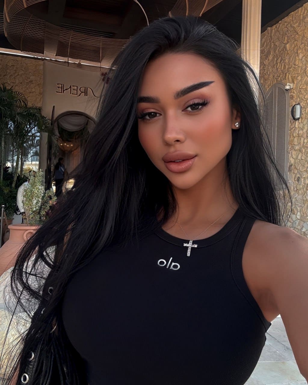 Inara with long dark hair in a black sleeveless top and cross necklace, posing outdoors near stone walls and plants.
