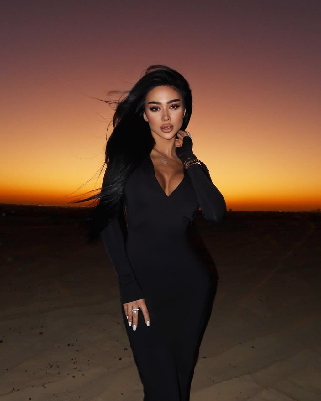 Woman in a long-sleeved black dress with deep V-neck stands in a desert at sunset, hand near her neck.