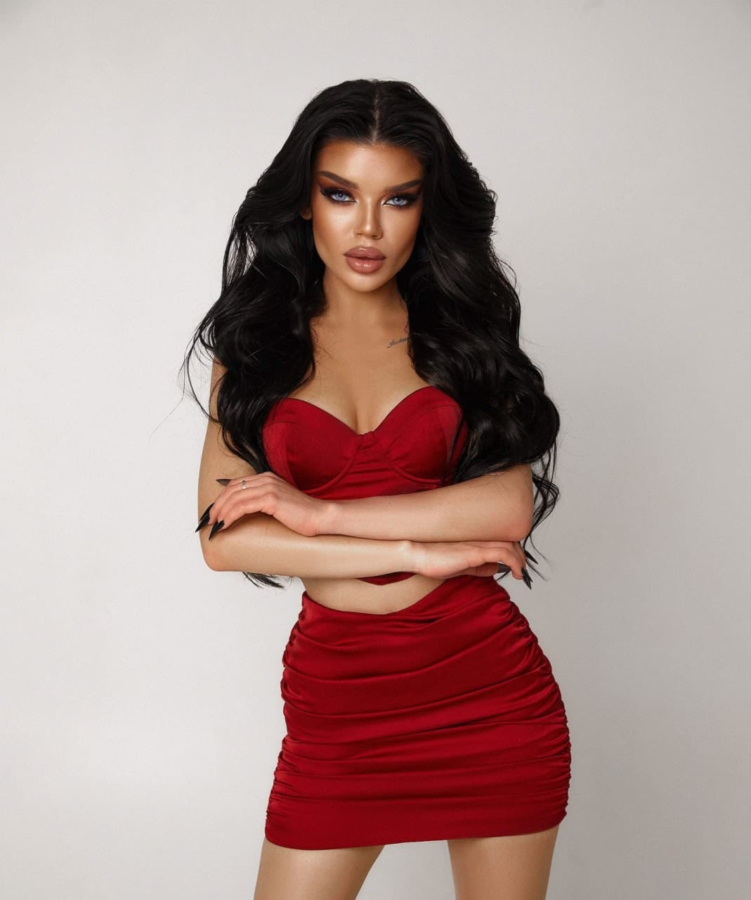 Jasmine stands with arms crossed, wearing a red satin strapless crop top and ruched mini skirt against a plain light background.