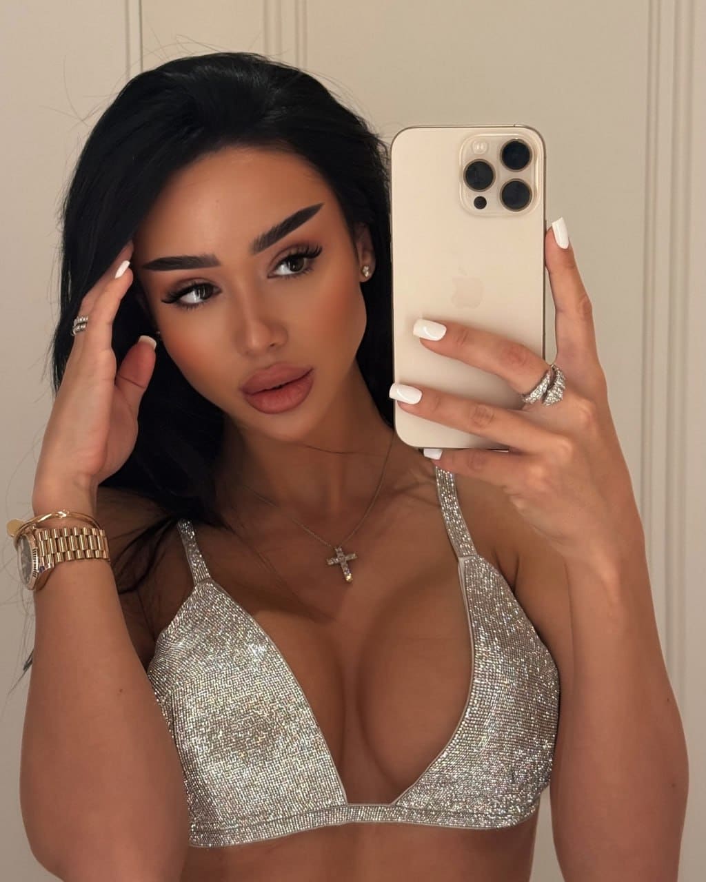 Mirror selfie of a woman in a silver sequined bra top and gold watch, one hand near her head, holding a phone.