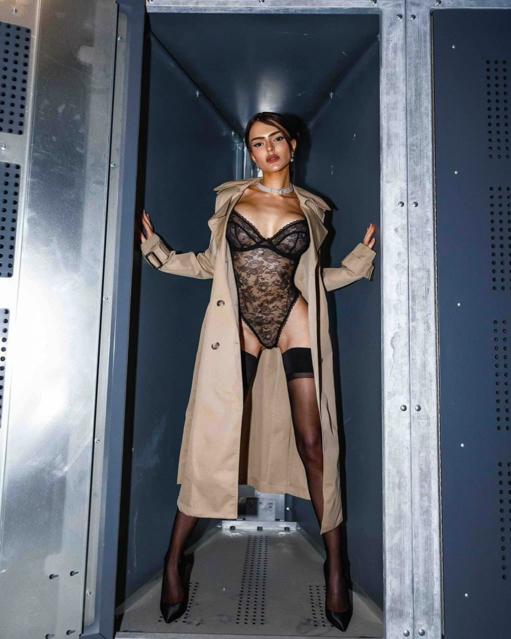 Woman stands in a metallic shaft wearing an open trench coat, black lace bodysuit, stockings, and heels, hands on walls.