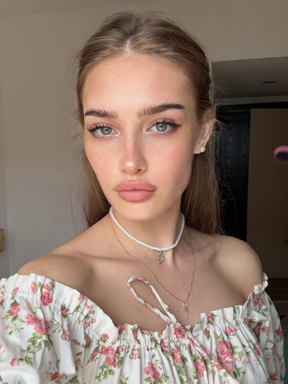 Esmeralda faces the camera indoors, wearing a white off-the-shoulder top with pink floral patterns, a pearl choker, and a gold necklace.