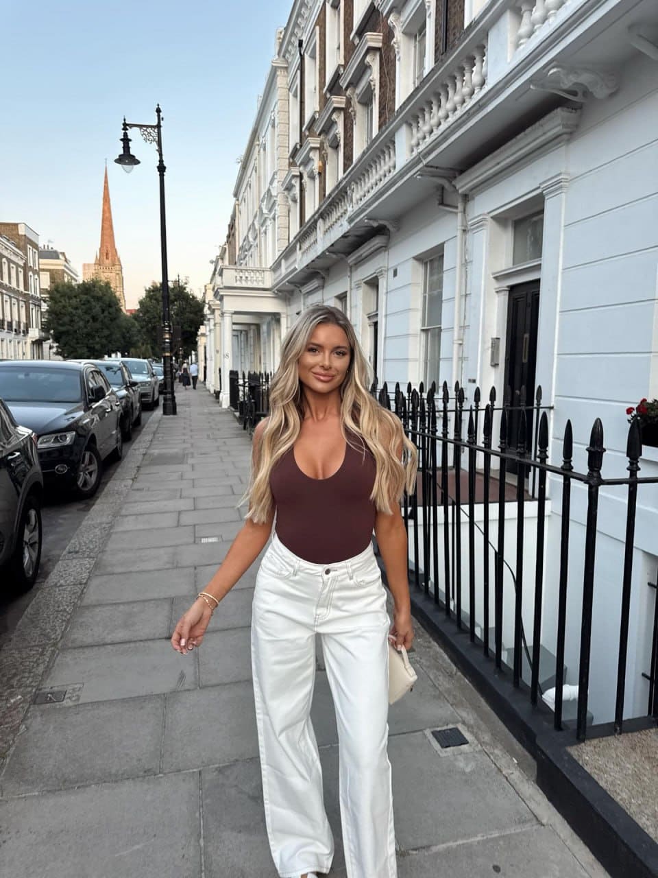 Angela walks a city sidewalk in a maroon sleeveless bodysuit and white high-waisted trousers.