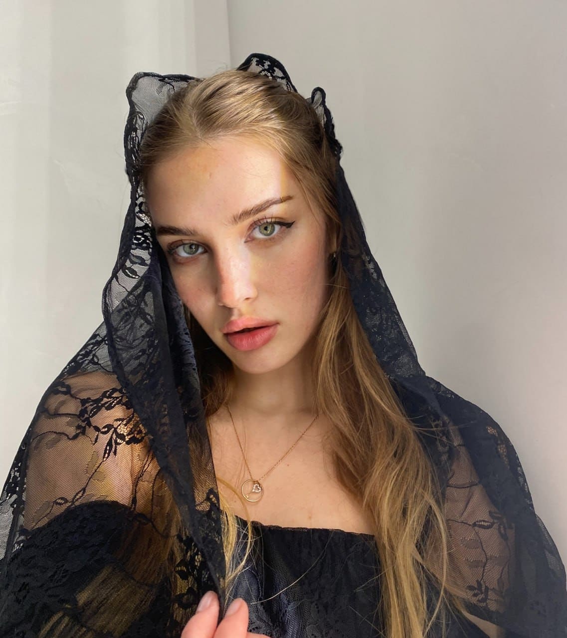 A blonde woman poses against a white wall wearing a black lace veil over her head and shoulders, a black strapless top, and a thin gold necklace.