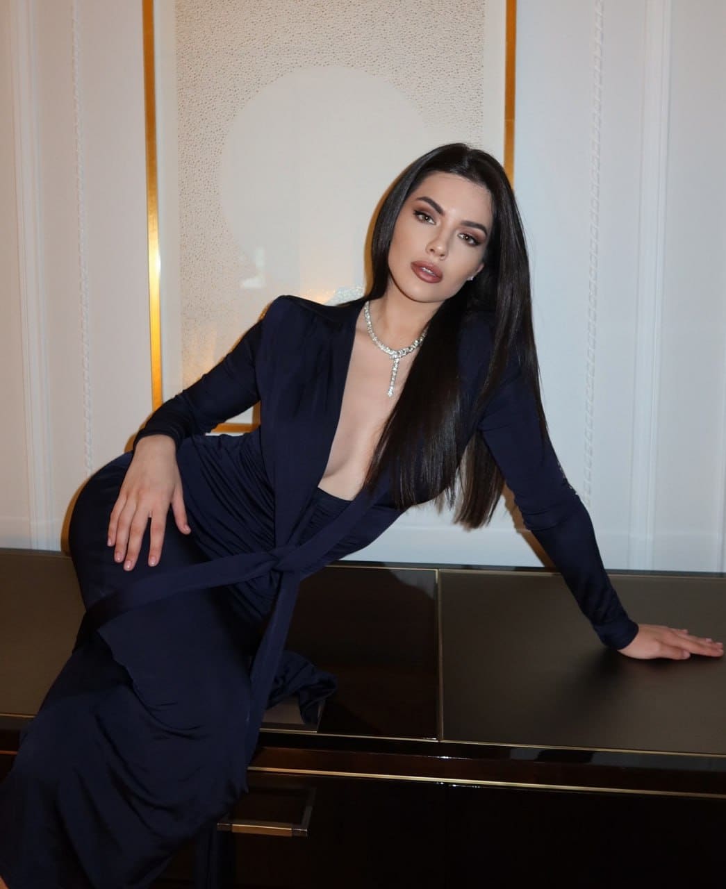 Lydia leans against a dark surface in a floor-length navy gown with long sleeves, a plunging neckline, and a diamond necklace.