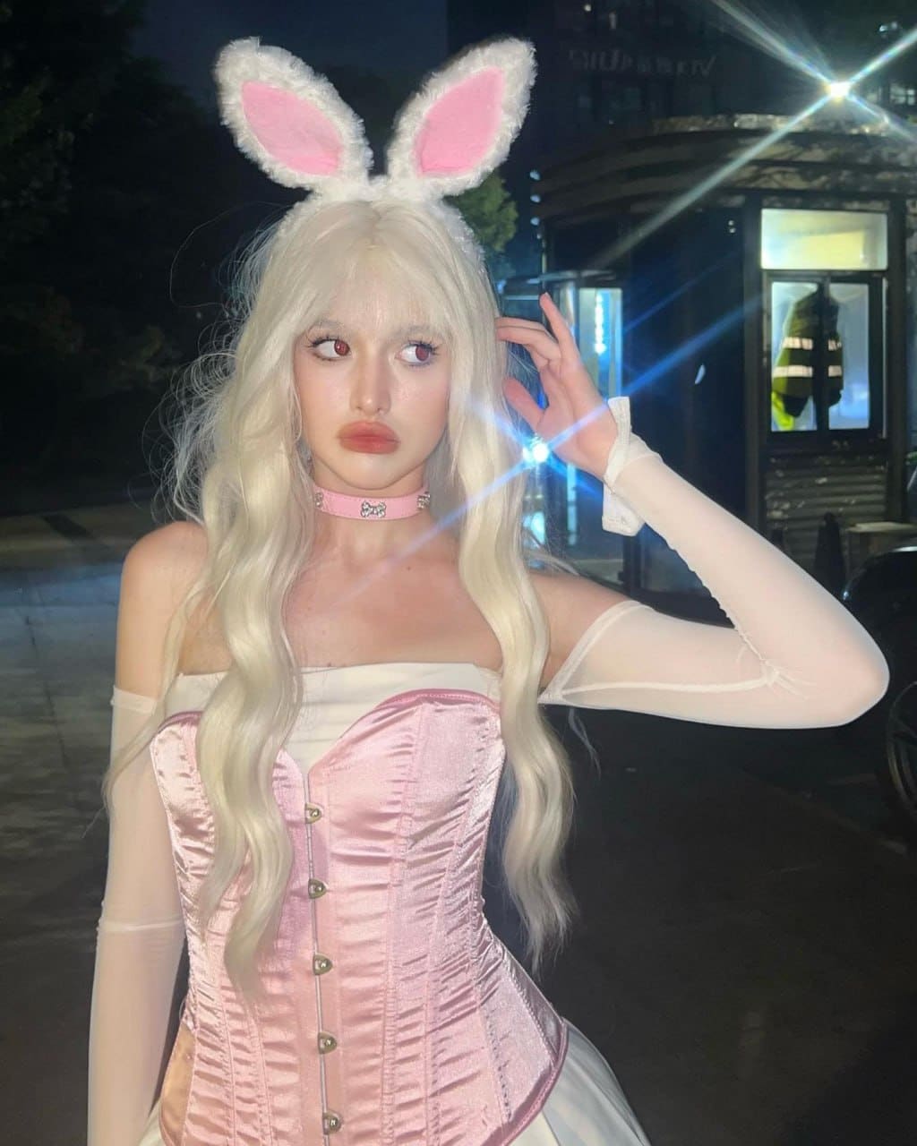 A person with platinum blonde hair and bunny ears wears a pink satin corset and sheer sleeves, posing outdoors at night.
