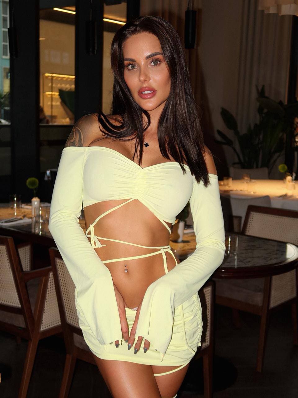 In a restaurant, a woman stands wearing a pale yellow off-the-shoulder top with detached sleeves and a matching mini skirt.