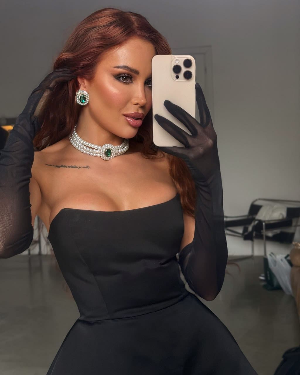 A mirror selfie shows a woman with auburn hair in a black strapless dress and sheer opera gloves, wearing a pearl choker and earrings.