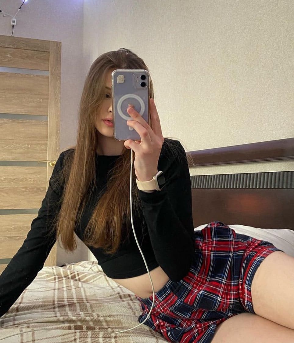Mirror selfie of a woman sitting on a bed in a black crop top and red plaid mini skirt, exposing midriff and legs.