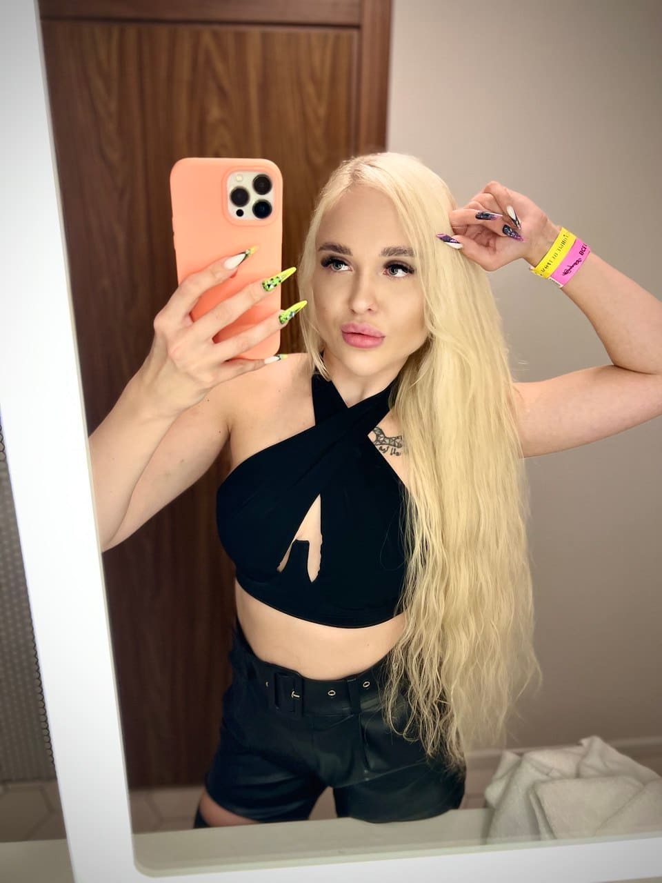 Mirror selfie of a woman in a black halter top with a chest cutout and black shorts, holding an orange-cased phone and touching her hair.