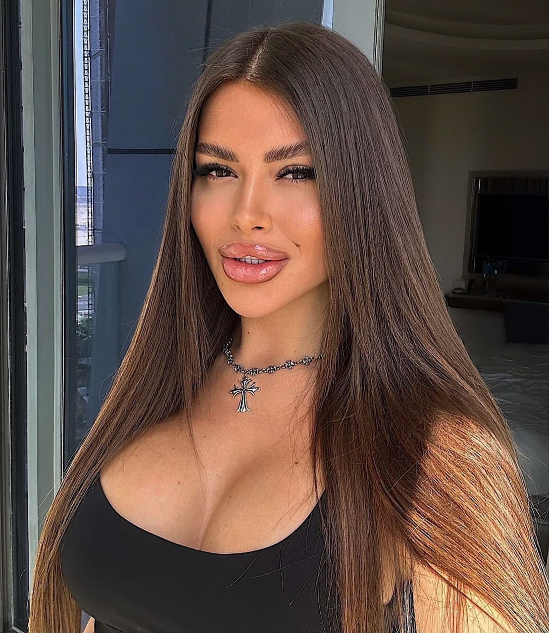 Woman with long dark hair poses indoors in a black top with a deep scoop neckline and silver cross necklace.
