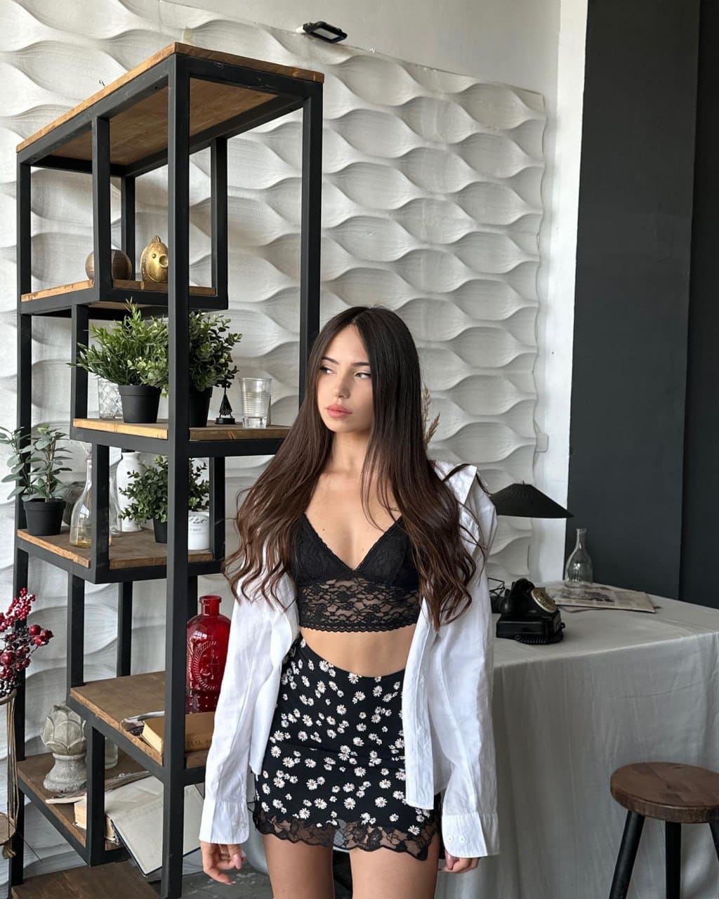 Josephine wears a black lace bralette, floral skirt, and open white shirt, standing by a black shelf and white wall.