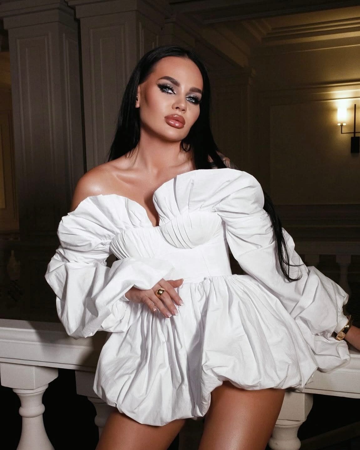 Arina leans against a white balustrade indoors, wearing a white off-the-shoulder mini dress with voluminous sleeves and a structured bodice.
