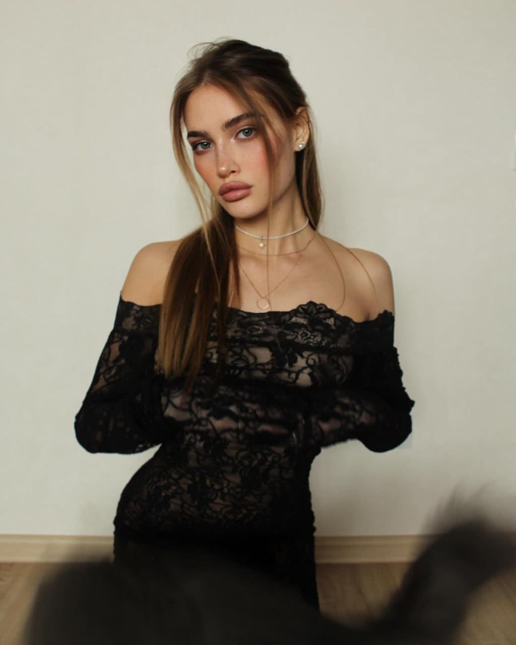 A woman with a side ponytail stands against a plain wall, wearing a sheer black lace off-the-shoulder top with long sleeves and layered necklaces.