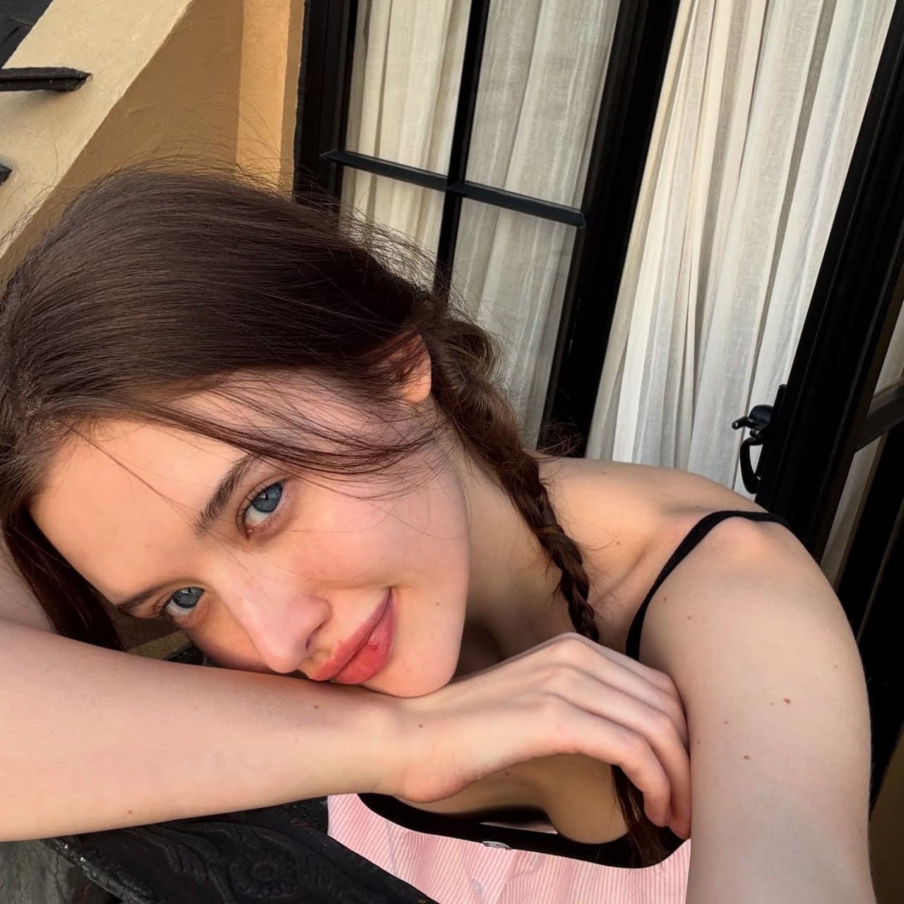 A woman with a loose braid rests her chin on crossed arms, wearing a pink and white striped top against sheer white curtains.
