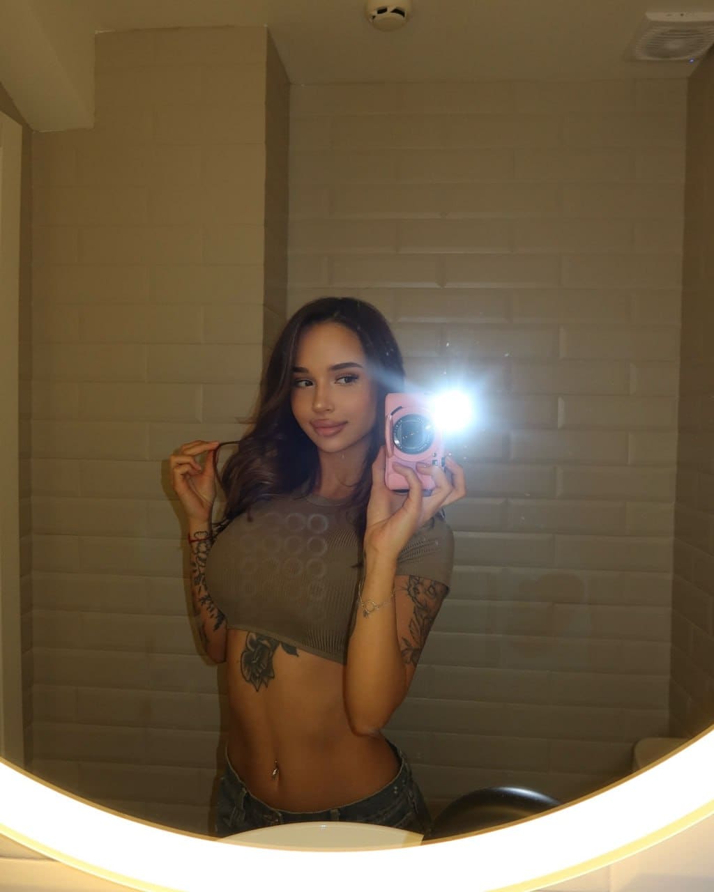A woman takes a mirror selfie in an olive green crop top and denim shorts, standing before white brick walls.