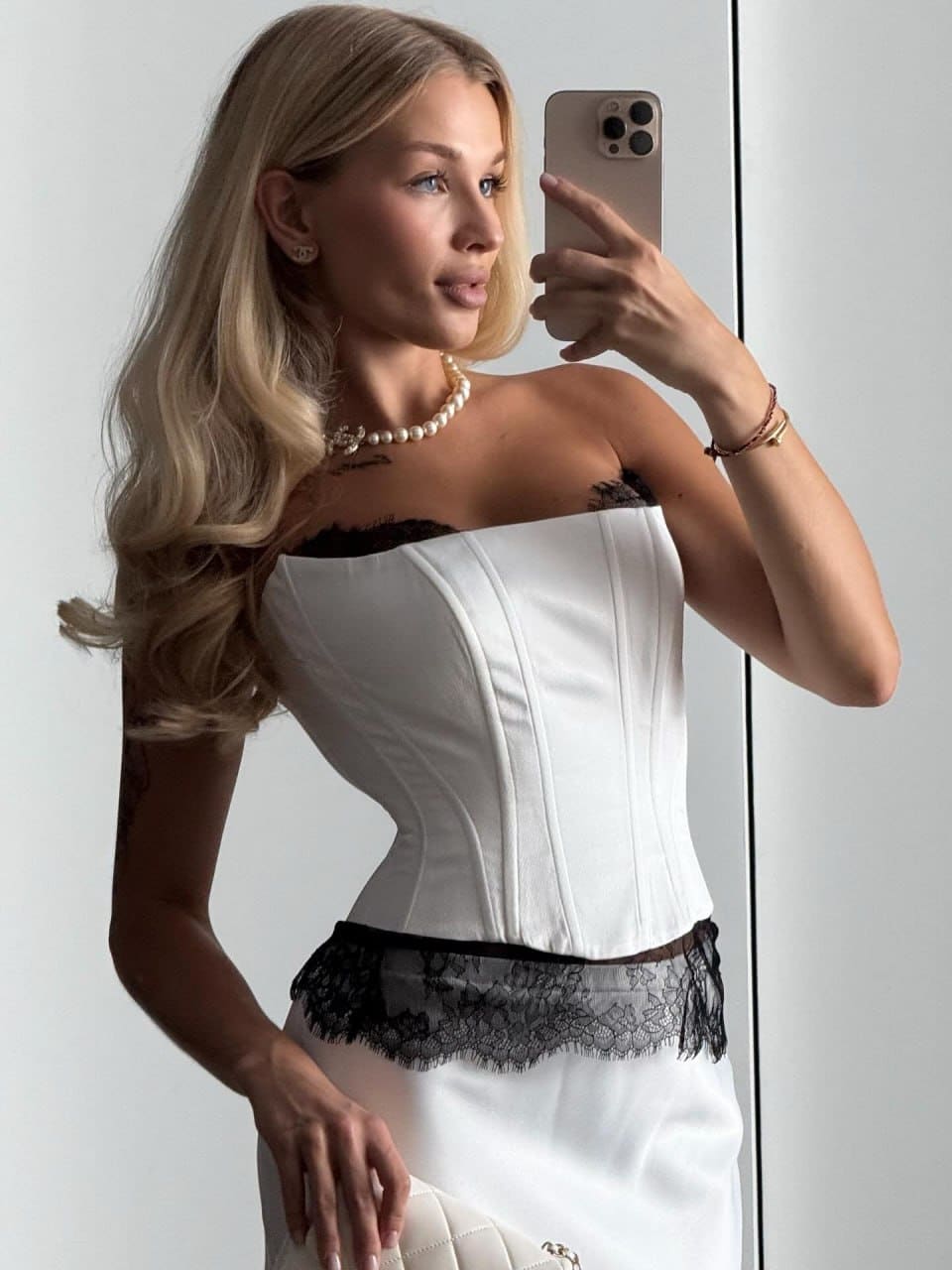 A mirror selfie shows a woman in a white strapless corset top with black lace trim, white skirt, and pearl necklace against a light wall.