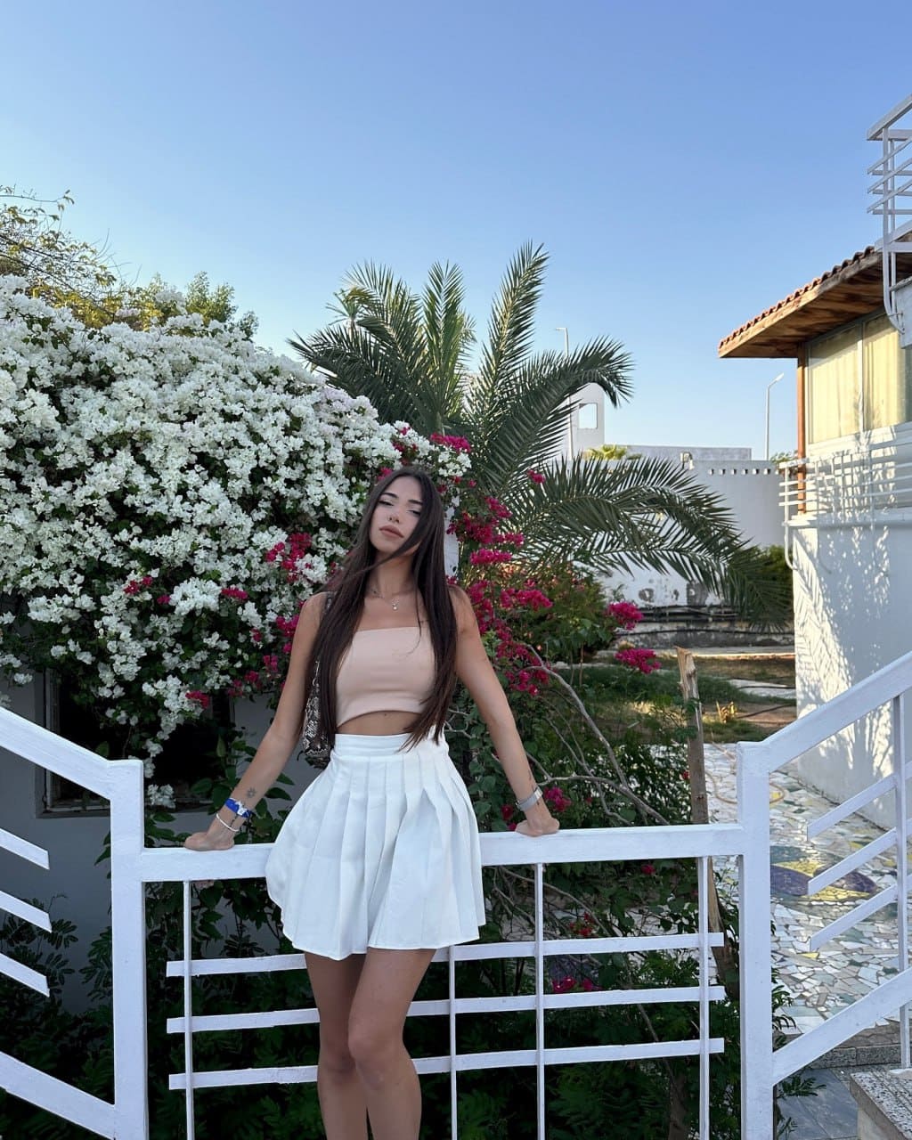 A woman leans back on white stairs in a beige tank and white skirt, framed by flowering bushes and palm trees.