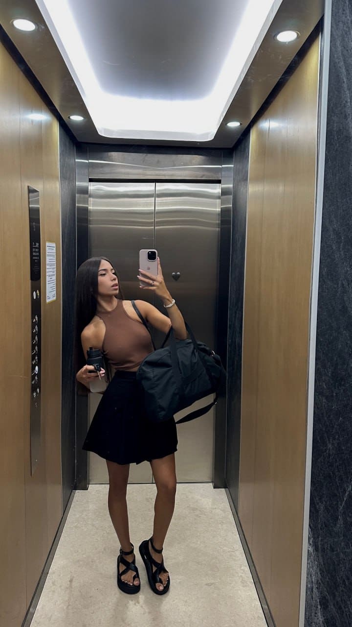 A woman takes an elevator mirror selfie in a brown cutout top, black skirt, and sandals, holding a phone and bag.