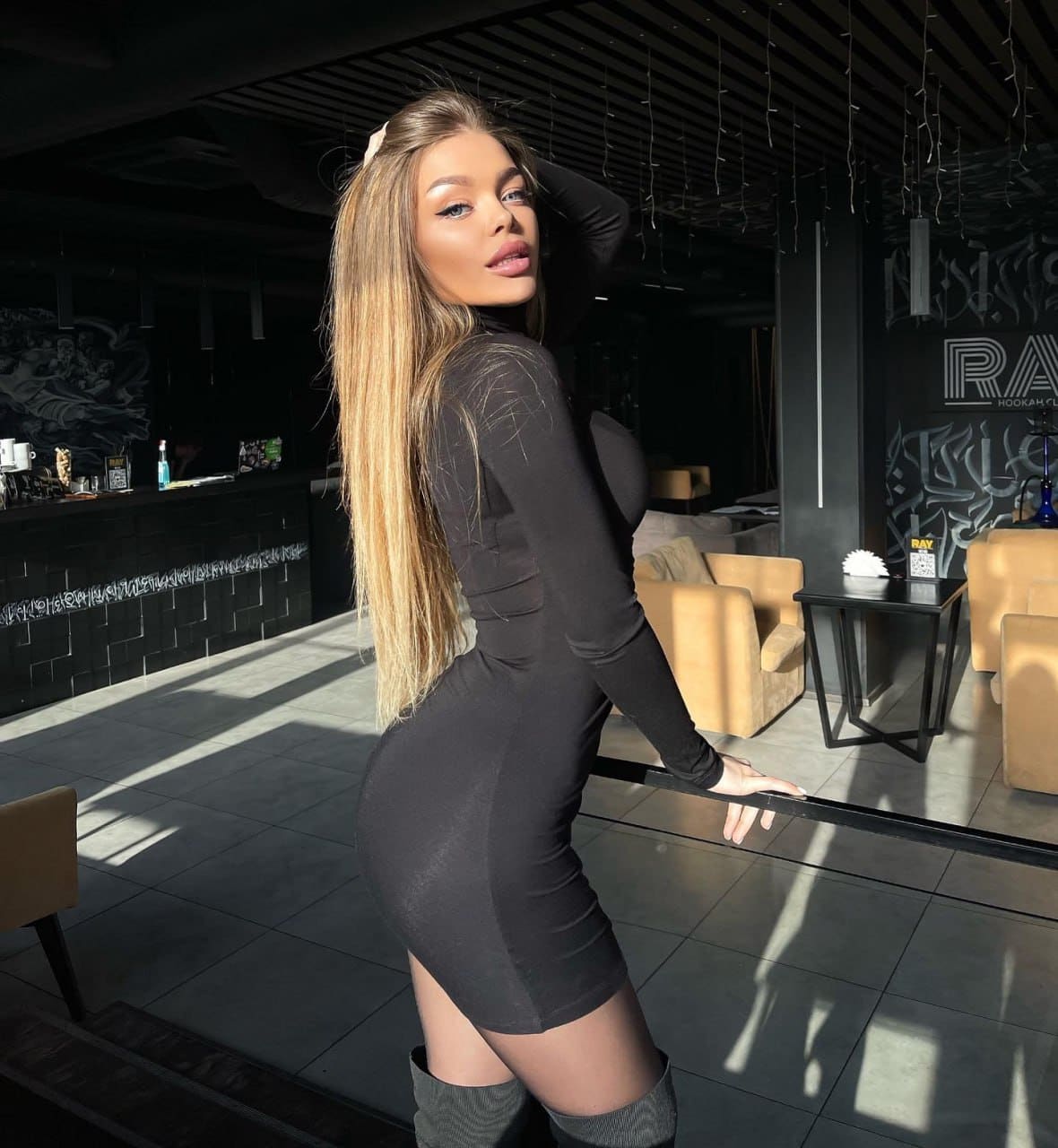A blonde woman stands in profile in a lounge, wearing a tight black mini dress and thigh-high boots, with one hand resting on her head.