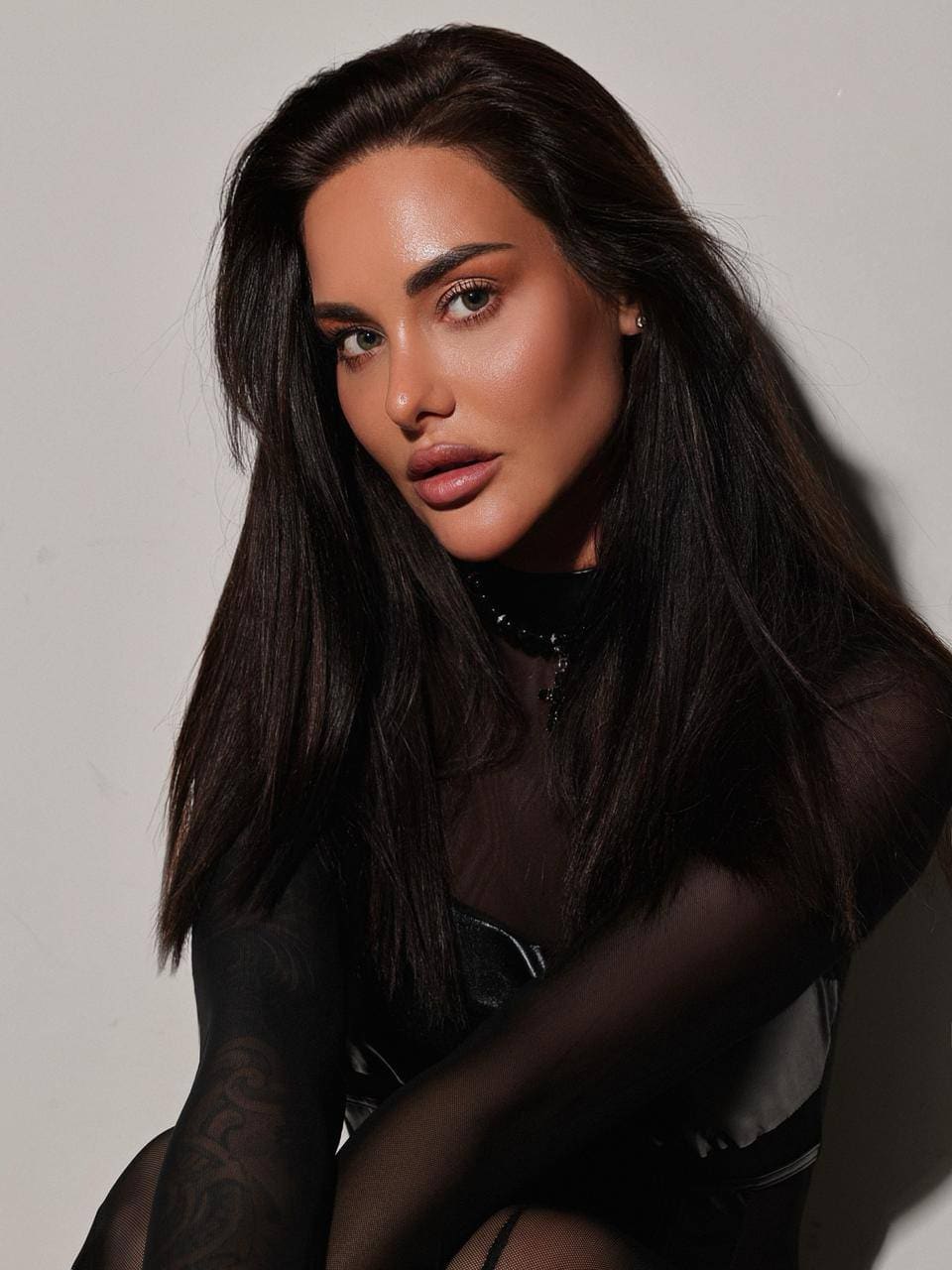 Eliana with long dark hair poses with arms crossed, wearing a sheer black mesh top over a dark undergarment against a plain background.