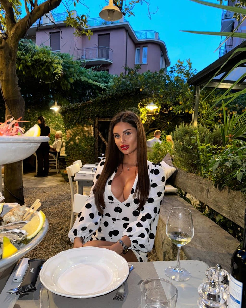 Alena sits at an outdoor table in a white polka-dot dress with a plunging neckline, a glass of white wine in the foreground.