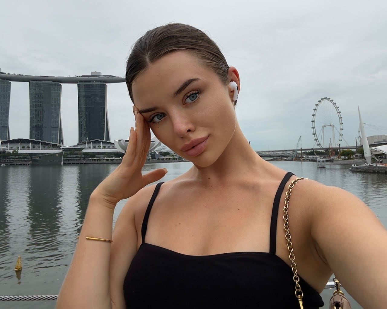 Emmeline poses outdoors with a hand to her temple, wearing a black top with gold straps against a waterfront with a hotel and Ferris wheel.