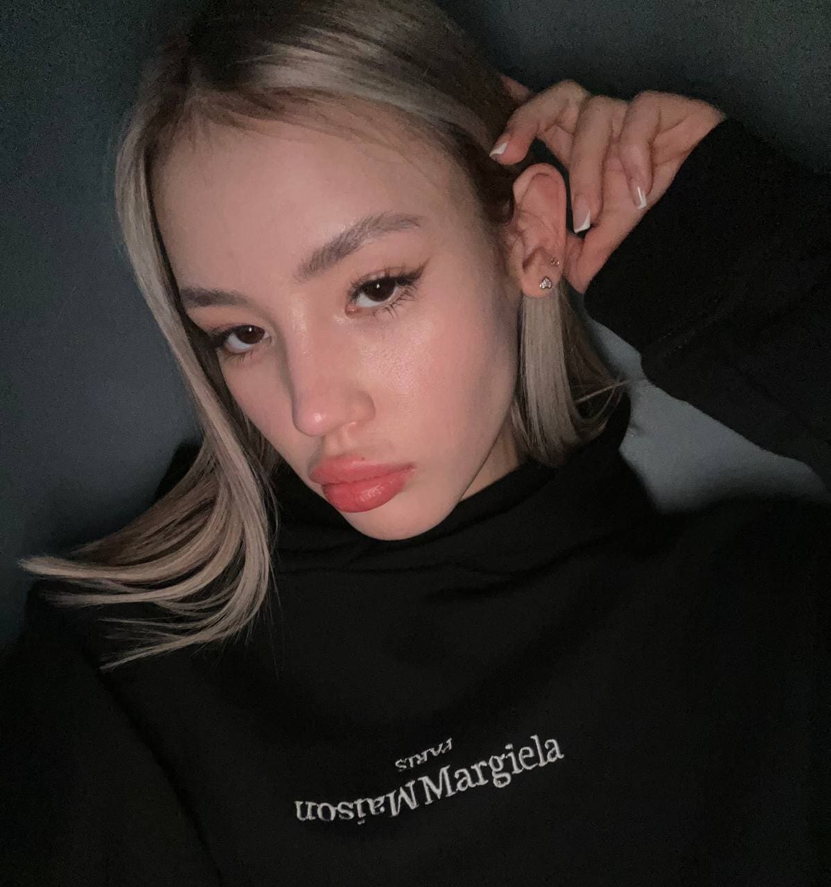 A close-up selfie of a blonde woman in a black hoodie with white text, raising a hand near her ear against a dark background.