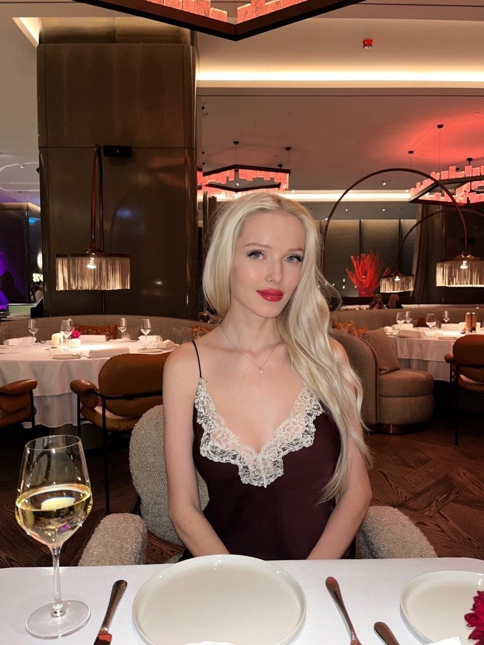 Nova sits at a restaurant table in a maroon slip dress with lace trim, a glass of white wine in the foreground.