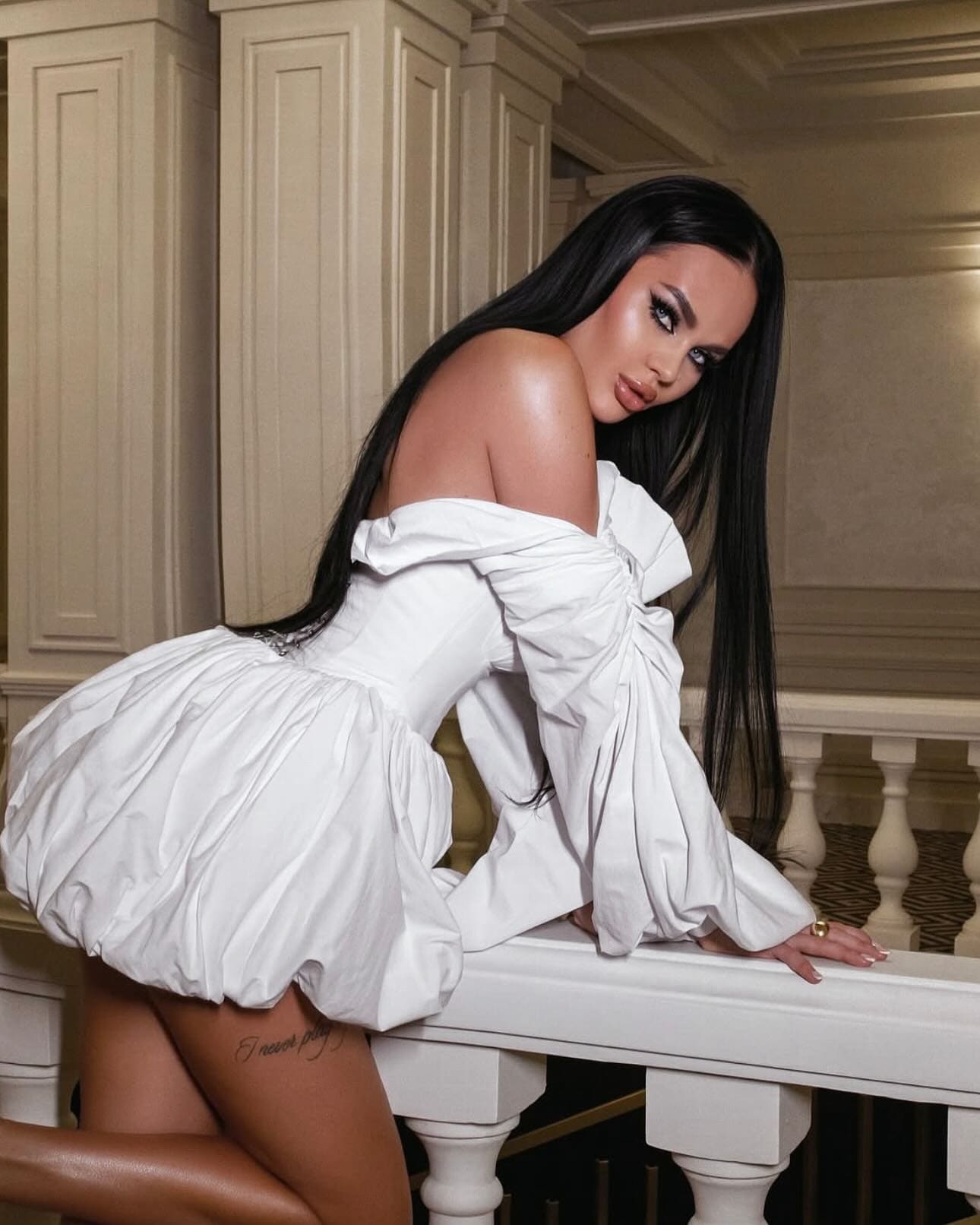 A woman leans forward on a white balustrade against cream paneled walls, wearing a white off-the-shoulder mini dress with a puffed skirt.