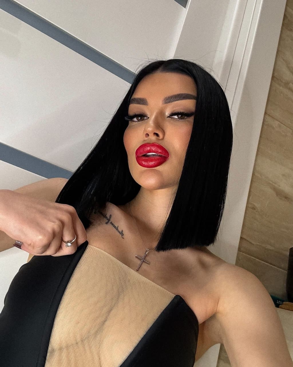 Wearing red lipstick and a strapless top with a sheer nude panel, she poses indoors near a white door with a cross necklace.