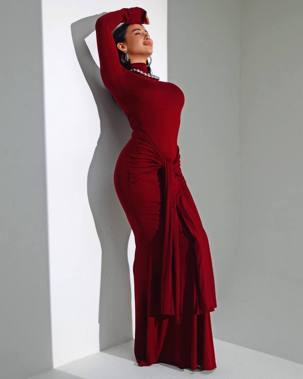 A woman stands in profile against a white wall, wearing a floor-length burgundy dress with long sleeves and a high neck.