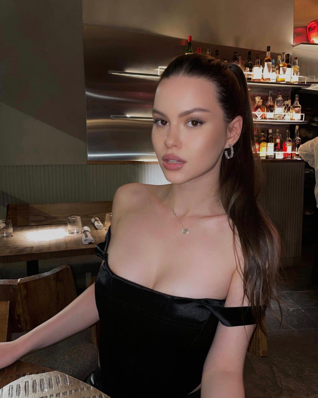 A woman with a high ponytail wears a black off-the-shoulder top while seated at a bar with shelves of liquor bottles behind her.