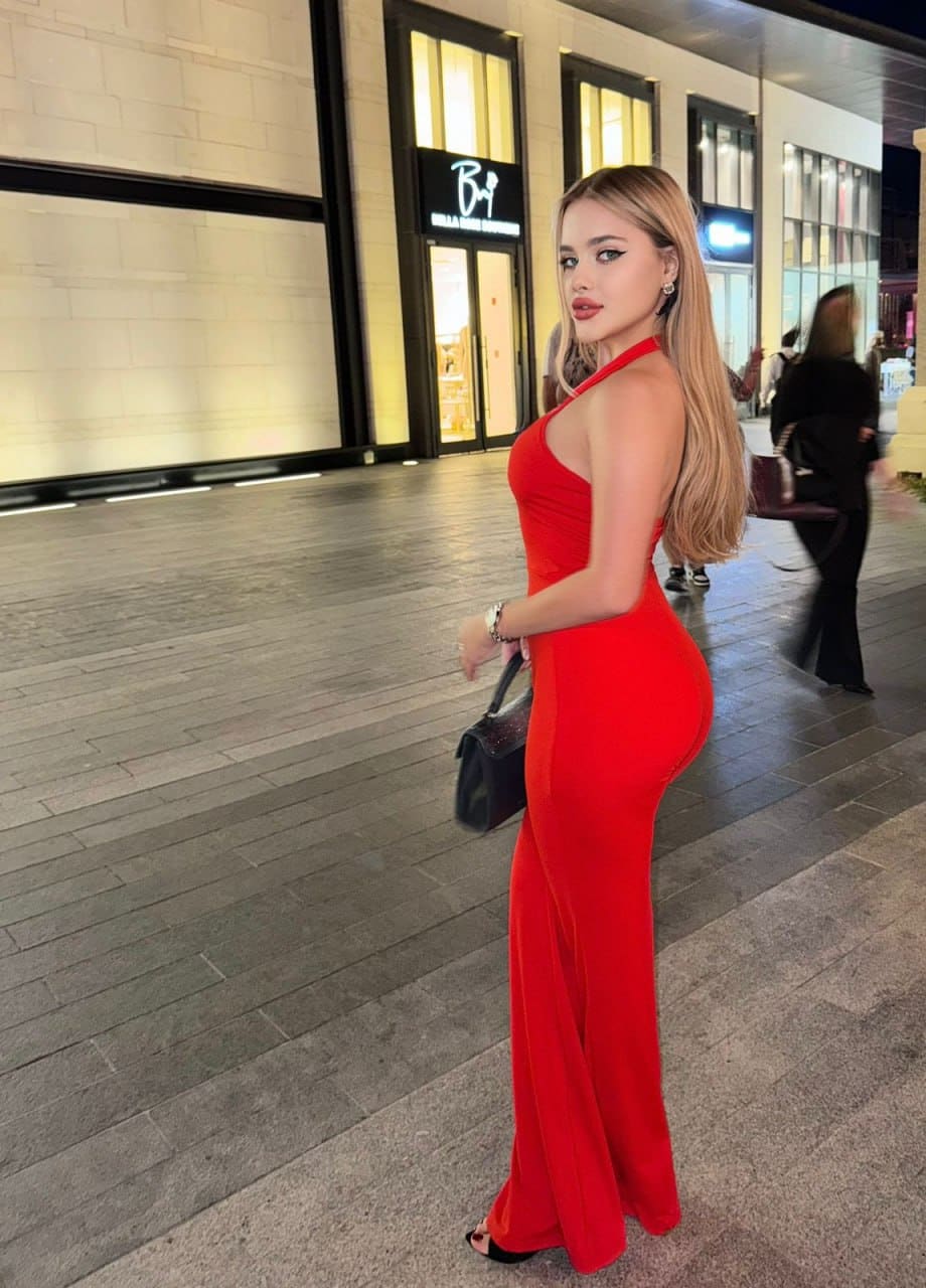 Standing in profile on a night walkway, a woman wears a red halter-neck maxi dress and holds a small black handbag.