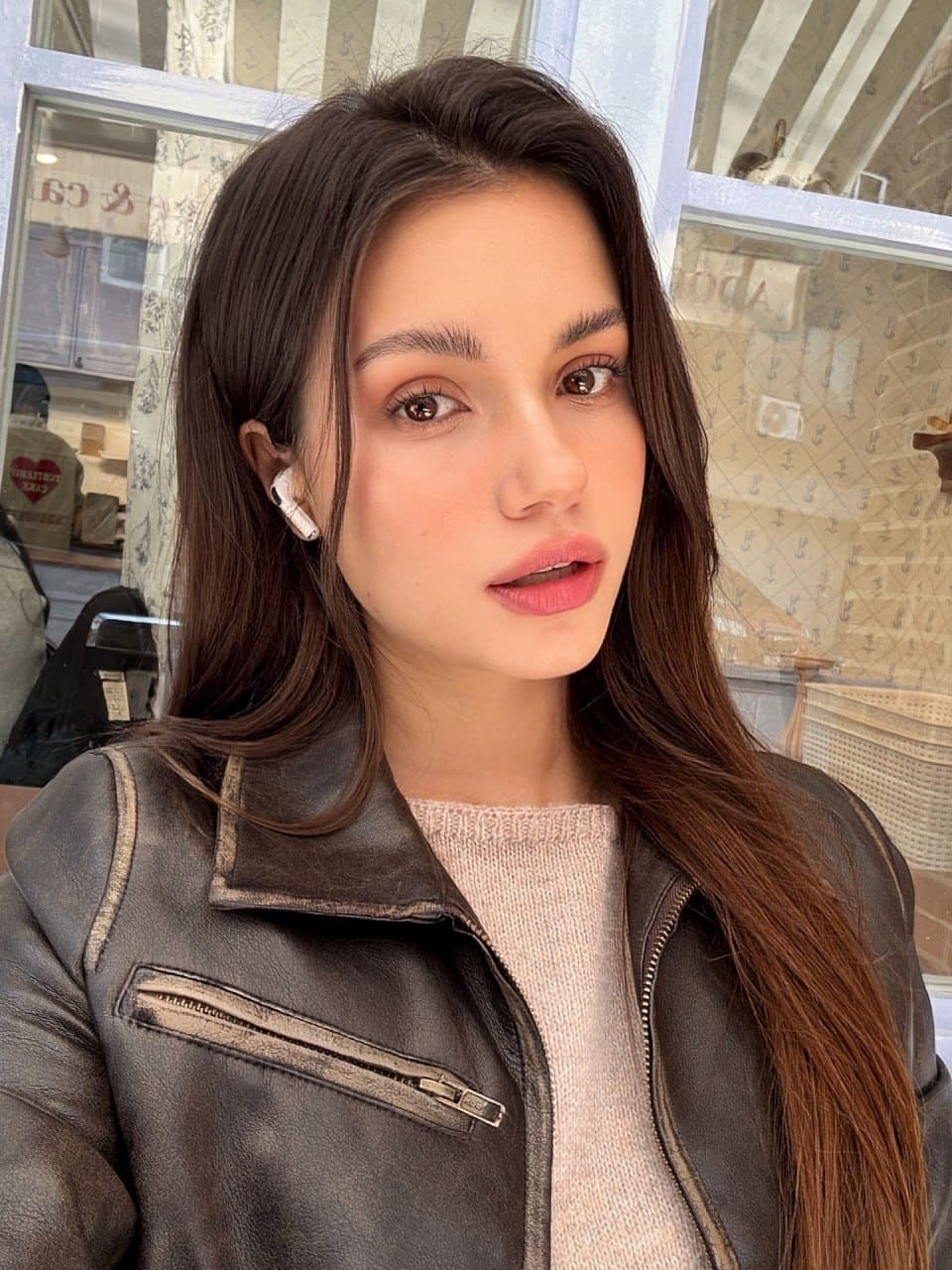 Mira wears a dark leather biker jacket over a pink sweater and white earbud, posing for a selfie in front of a windowed storefront.