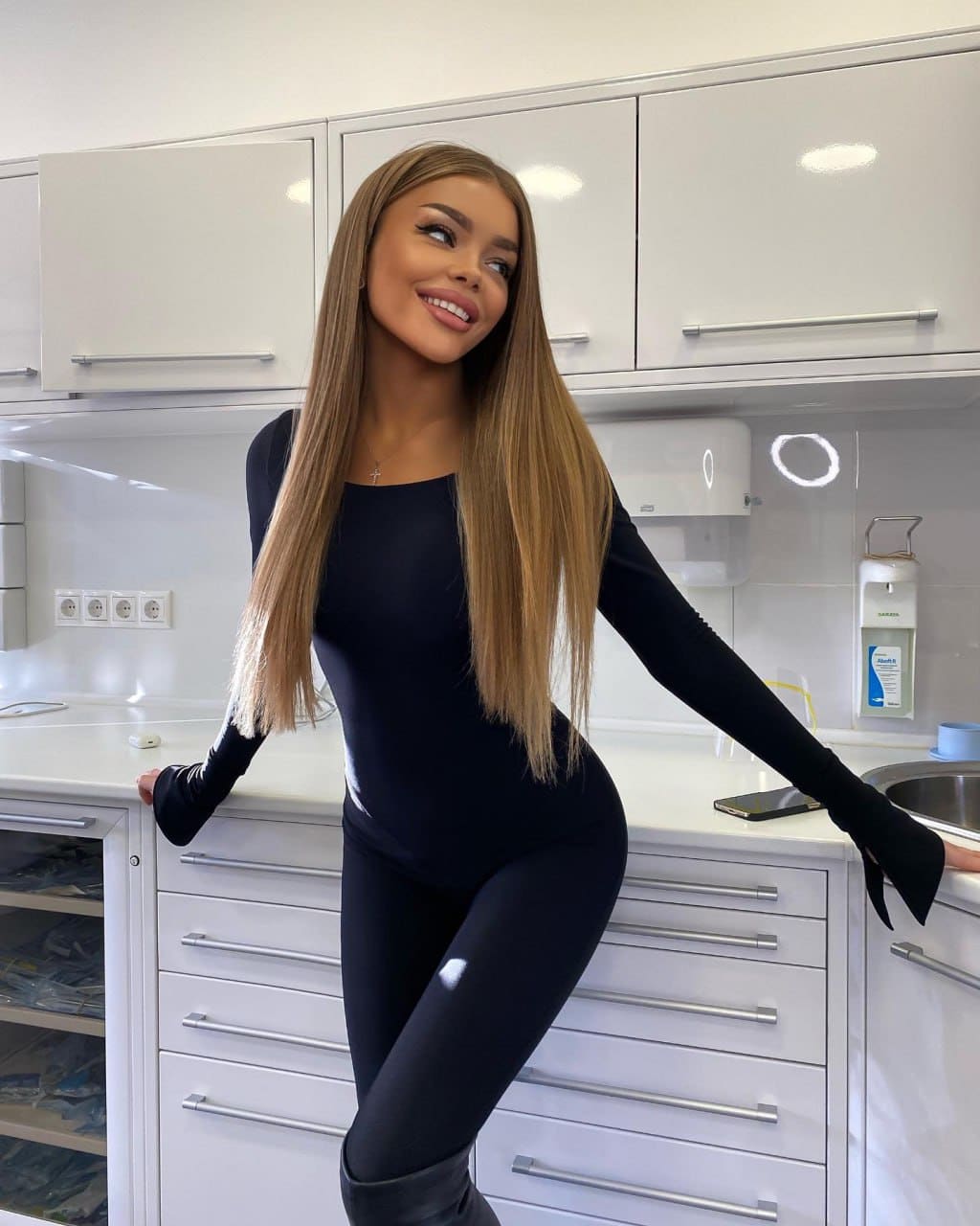 Halia stands in a white kitchen wearing a black long-sleeved bodysuit, leggings, and tall boots, leaning back against a counter with arms extended.