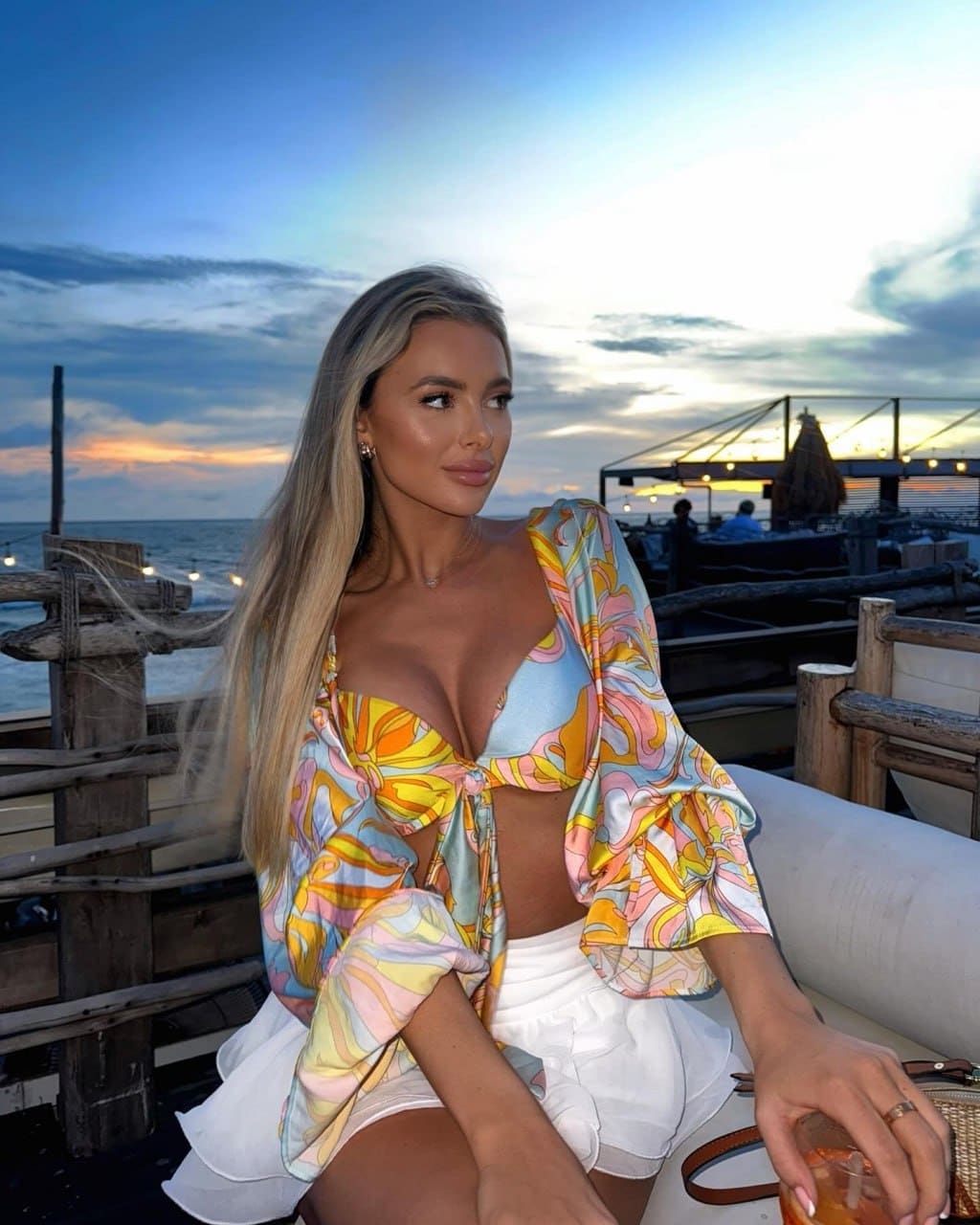 Blonde woman sitting outdoors at dusk by a railing, wearing a patterned top and white skirt, holding a drink.