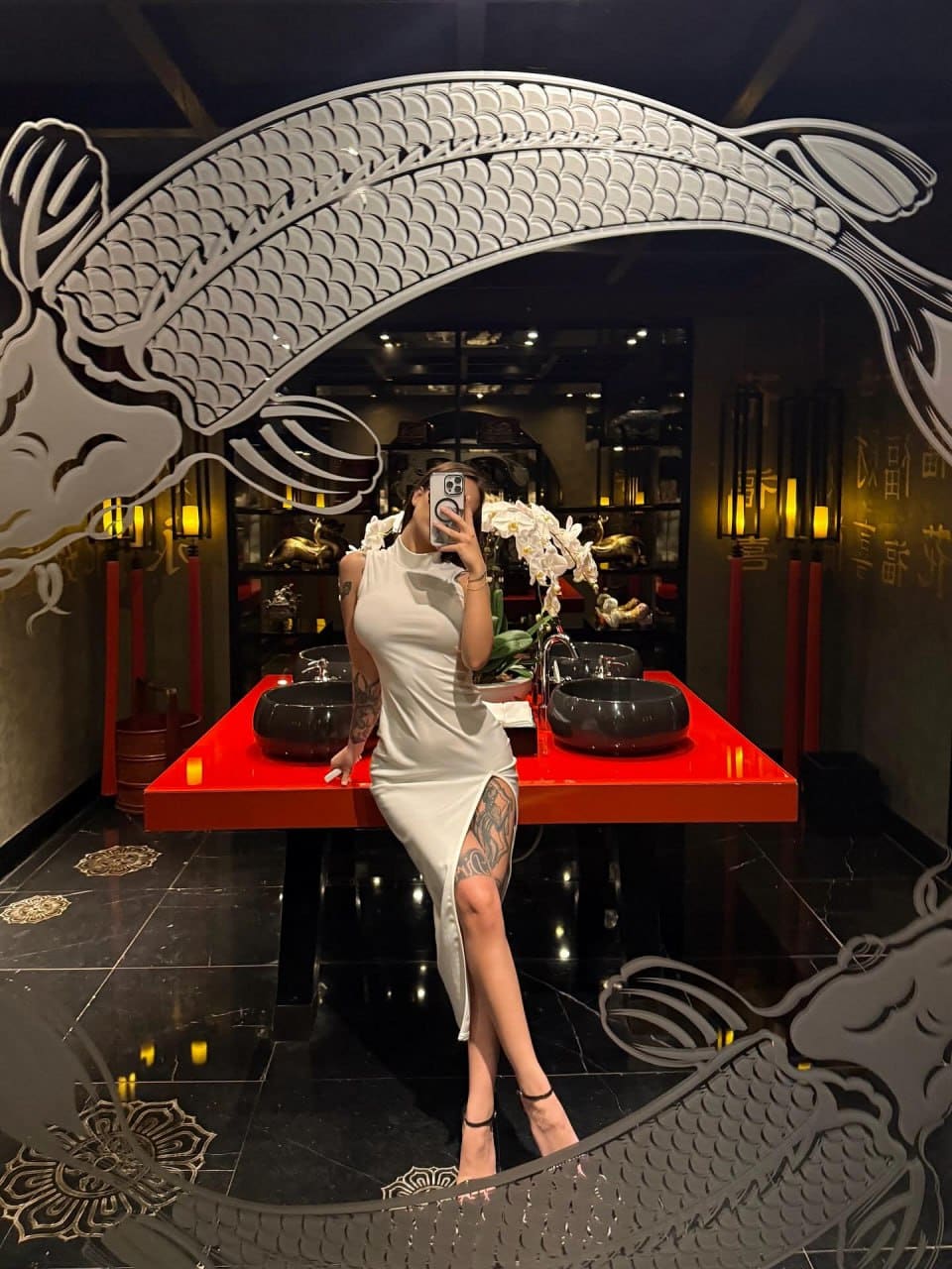 A woman leans against a red table in a mirror selfie, wearing a white high-neck dress with a thigh slit and strappy heels.