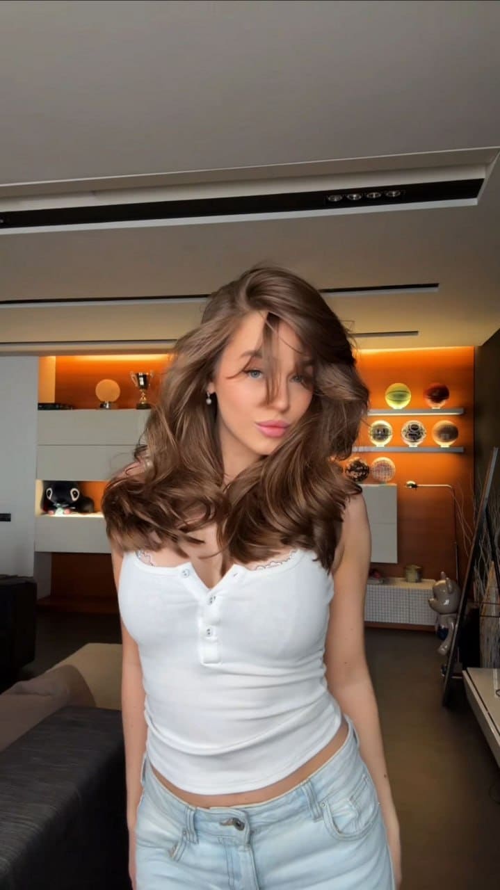 A woman tilts her head slightly while wearing a white buttoned henley top and light blue jeans, standing in a room with orange accent lighting.