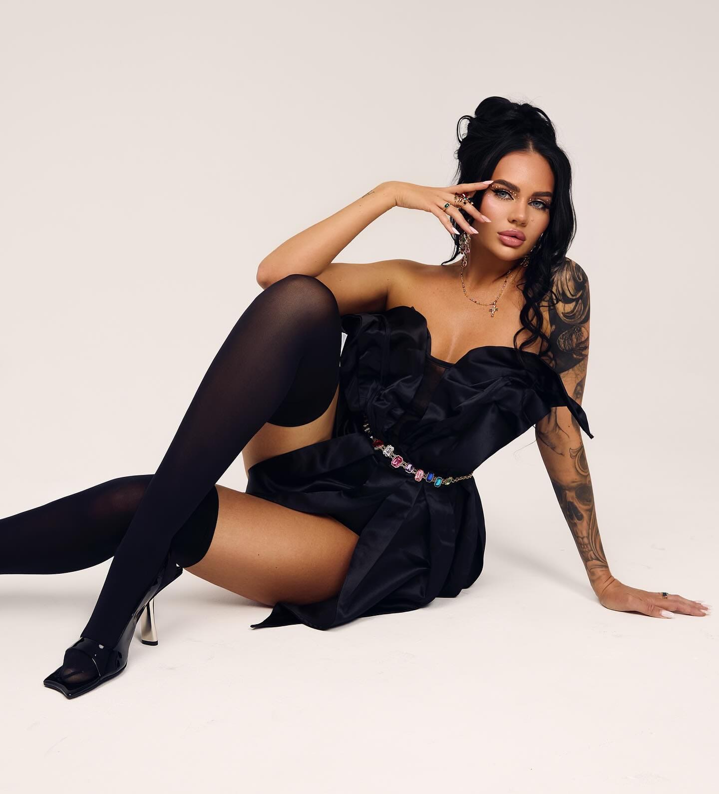 A woman with a high ponytail sits on a white studio floor in a strapless black satin dress, sheer stockings, and black sandals.