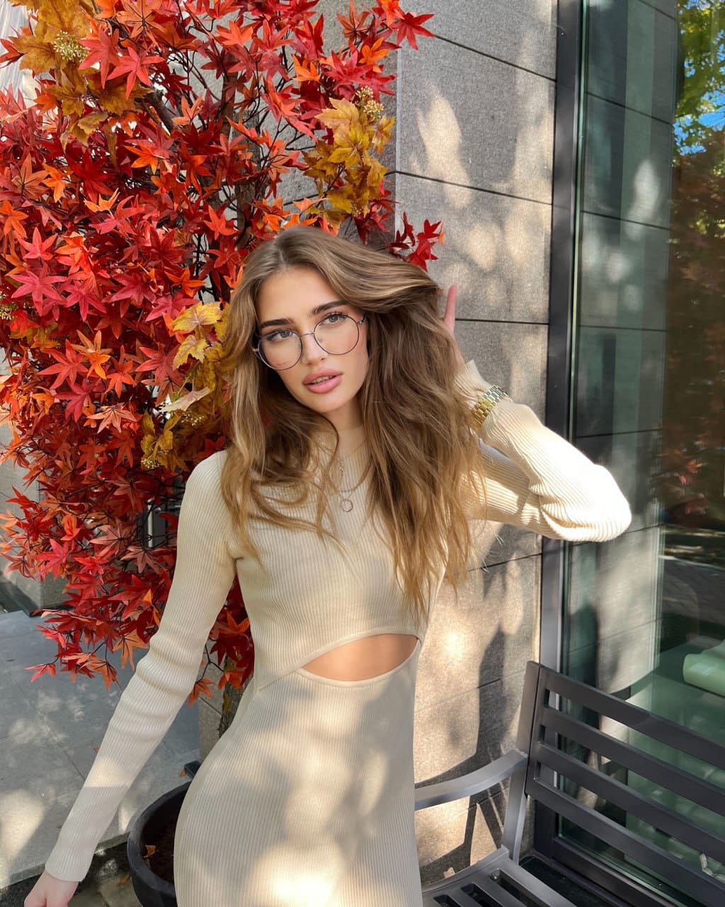 A woman with glasses and wavy hair stands outdoors by a tree with red leaves, wearing a cream ribbed knit dress with a midriff cutout.
