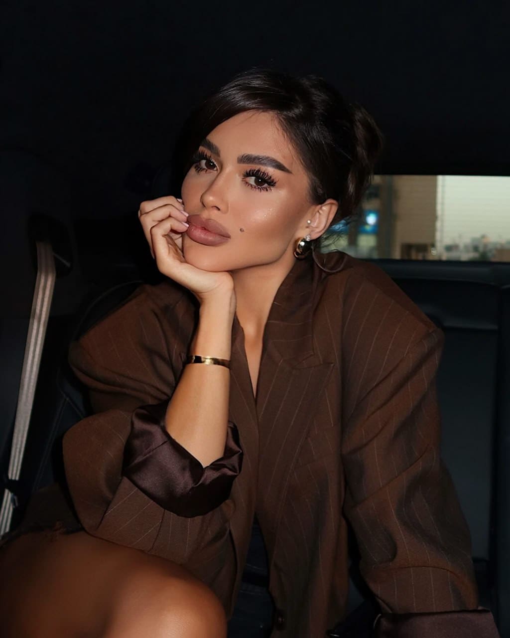 Kiki sits in a vehicle with chin on hand, wearing a brown pinstripe blazer with satin cuffs and hoop earrings.
